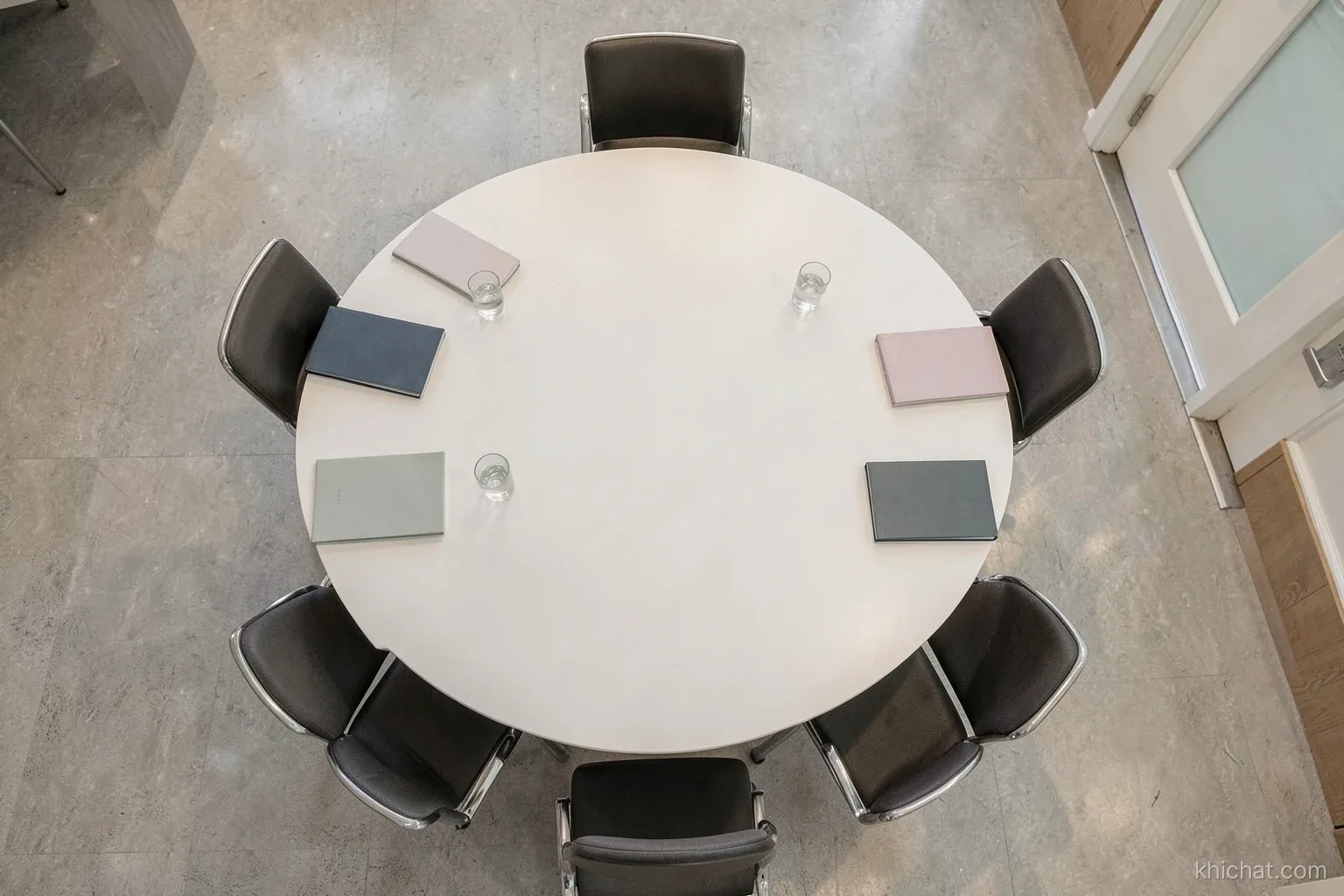 Top-down view of an empty oval meeting table with neatly arranged chairs, water glasses and closed notebooks, one chair at the far end facing the door, soft overhead light